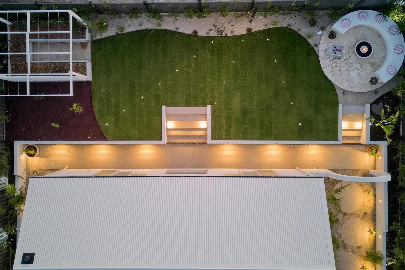 Overhead view of a completed backyard at dusk, with a curved lawn, a circular firepit seating area in crazy paving, and a lit outdoor kitchen along the boundary
